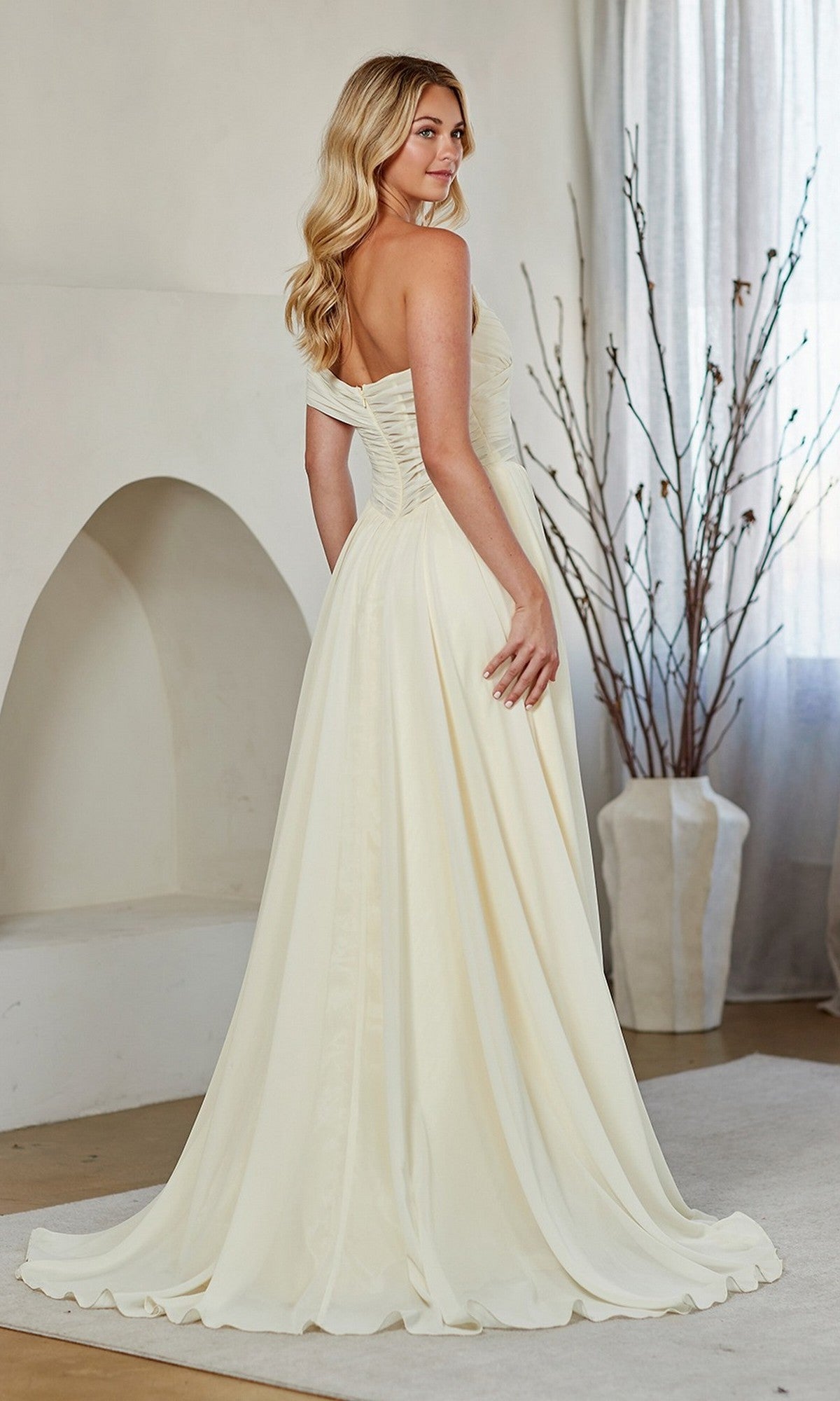 Woman wearing the Amelia Couture 9097, a long ivory off-the-shoulder prom dress with a fitted ruched bodice and flowing A-line skirt, stands indoors beside a vase with branches.